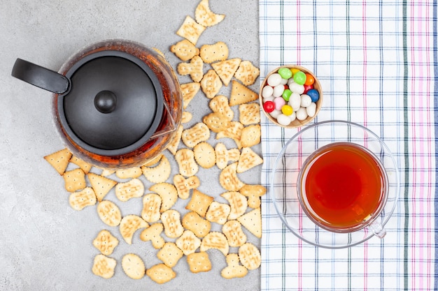 Teiera e una tazza di tè su un asciugamano con scaglie di biscotto sparse e una ciotola di caramelle sulla superficie di marmo.