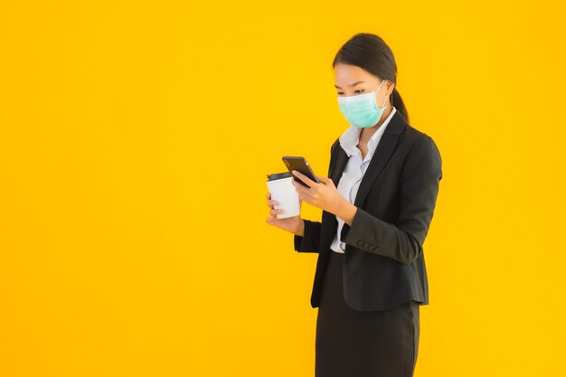 Telefono cellulare asiatico di uso della maschera di usura della donna di bei giovani affari del ritratto con caffè