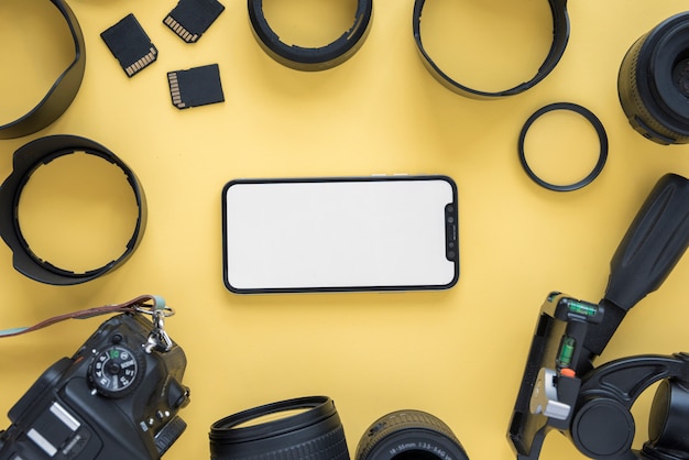 Telefono cellulare con schermo vuoto circondato da moderni accessori della fotocamera su sfondo giallo