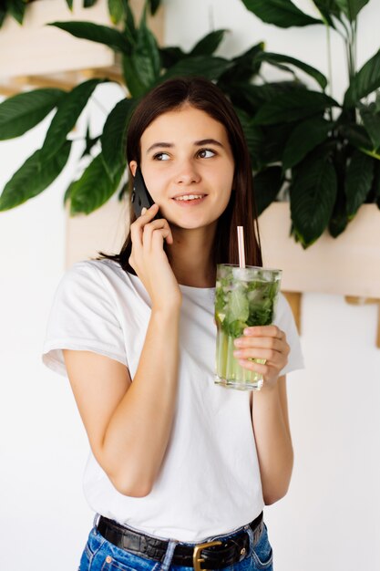 Telefono cellulare di conversazione della giovane ragazza graziosa e frullato bevente mentre stando alla tavola in un caffè