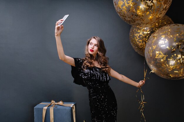 Tempo di festa di giovane donna attraente in abito di lusso nero, con lunghi capelli ricci bruna che fa selfie con grandi palloncini pieni di orpelli dorati. Presenta, festeggia, moderno.