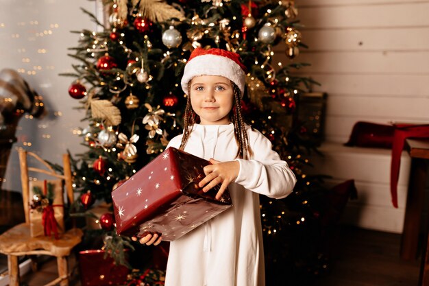 Tempo di Natale, bambino allegro in un cappello di Natale con il regalo di Natale che posa sopra l'albero di Natale