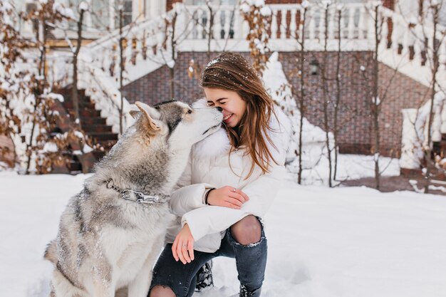 Tempo di nevicata invernale sulla strada del simpatico cane husky che bacia affascinante gioiosa giovane donna. Momenti belli, vera amicizia, animali domestici, vere emozioni positive.