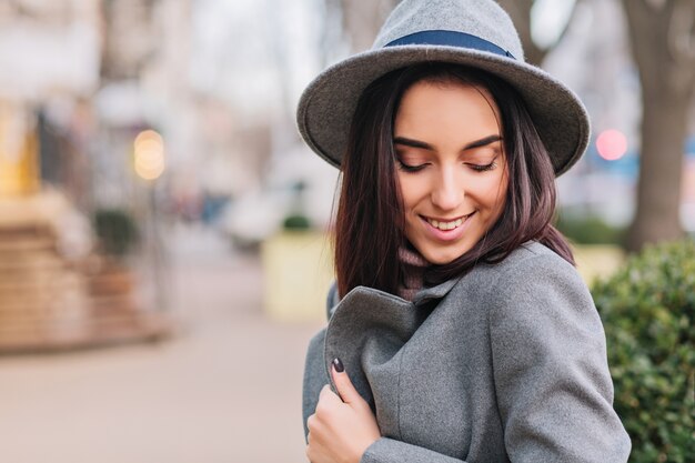 Tempo di passeggiata della città di affascinante giovane donna alla moda in cappotto grigio, cappello che cammina sulla strada in città. Sorridere con gli occhi chiusi, esprimendo le vere emozioni positive del viso, uno stile di vita di lusso, una prospettiva elegante.