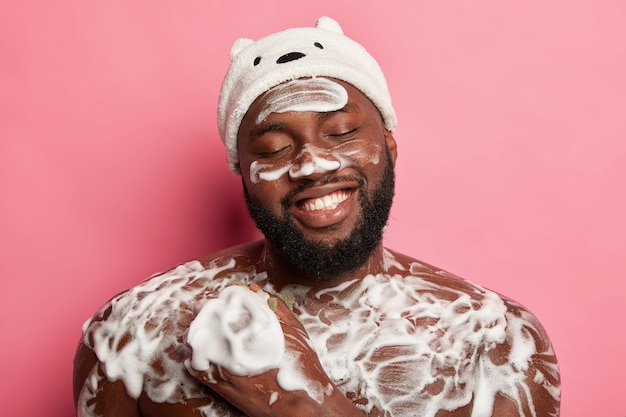 Tempo di relax e concetto di doccia. Felice l'uomo afro chiude gli occhi con piacere, sorride ampiamente, si lava con una spugna in bagno, applica gel schiumogeno