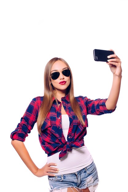 Tempo di selfie. Attraente giovane donna in abbigliamento casual che fa selfie dal suo smart phone e sorridente mentre in piedi contro il muro grigio