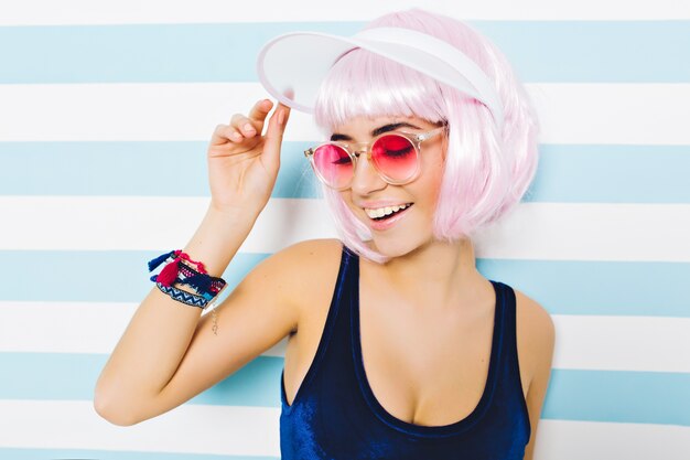 Tempo di spiaggia calda estate del ritratto del primo piano della donna alla moda in occhiali da sole rosa, con l'acconciatura rosa tagliata divertendosi sulla parete a strisce. Look sexy alla moda, umore allegro, sorridente con gli occhi chiusi.