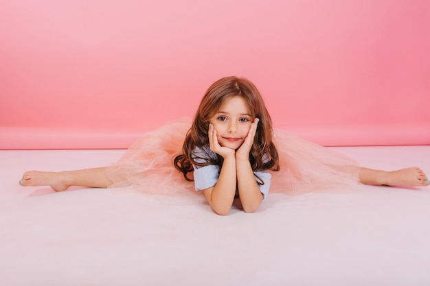 Tempo libero di gioiosa bambina affascinante facendo ginnastica divisa sul pavimento su sfondo rosa. Bambino carino elastico in gonna di tulle con lunghi capelli castani che sorride alla telecamera, esprimendo un umore allegro