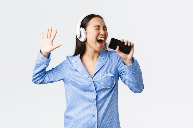 Tempo libero domestico, fine settimana e concetto di stile di vita. Ragazza asiatica eccitata e spensierata in pigiama, che suona l'app karaoke sullo smartphone, canta una canzone nel telefono cellulare mentre indossa le cuffie, sfondo bianco.