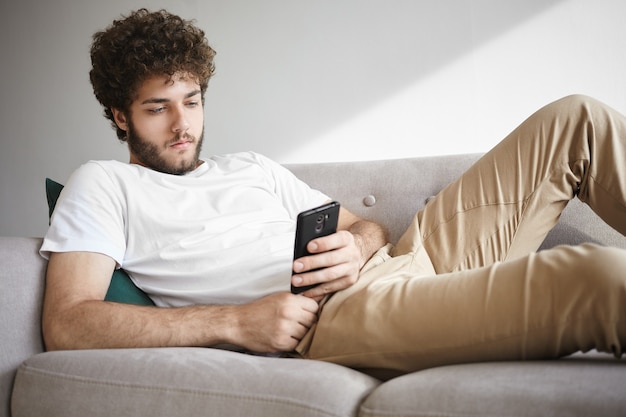 Tempo libero, tecnologia e concetto di comunicazione. Fantastico ragazzo con la barba lunga con una voluminosa acconciatura ondulata sdraiato sul divano nel suo appartamento moderno che ordina la consegna della pizza online sul suo dispositivo elettronico