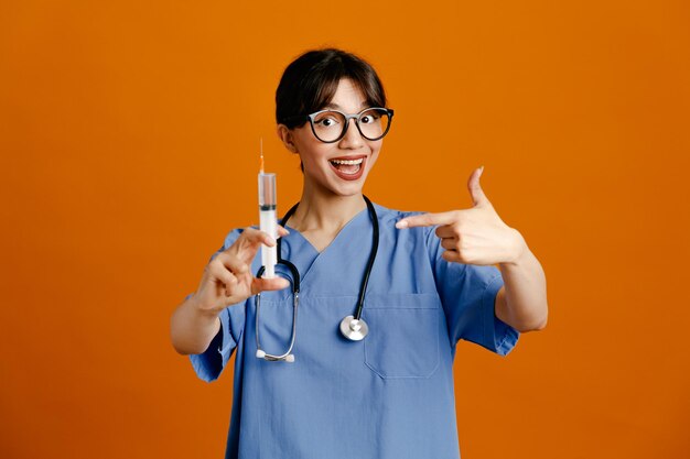 Tenendo e punta alla siringa giovane dottoressa che indossa uno stetoscopio fitth uniforme isolato su sfondo arancione