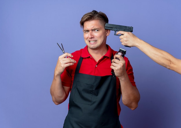 Tenendo la pistola al tempio del giovane barbiere maschio biondo ansioso in uniforme che tiene le forbici e il tagliatore di capelli isolato sullo spazio viola con lo spazio della copia