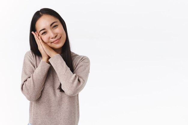 Tenera affascinante donna asiatica dai capelli scuri in maglione invernale, inclina la testa sciocca e carina, appoggiata sui palmi e contemplando qualcosa di affascinante con affetto, toccata da una scena adorabile, muro bianco