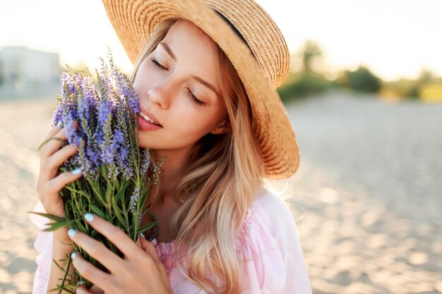 Tenera bella donna in cappello di paglia in posa sulla spiaggia soleggiata vicino all'oceano con bouquet di fiori. Close up ritratto.