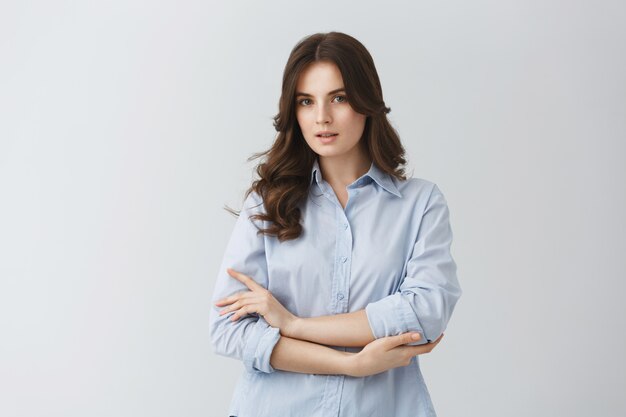 Tenera bella giovane donna con i capelli ondulati scuri in camicia blu con uno sguardo serio, in posa per una foto nell'articolo sulle giovani famiglie.