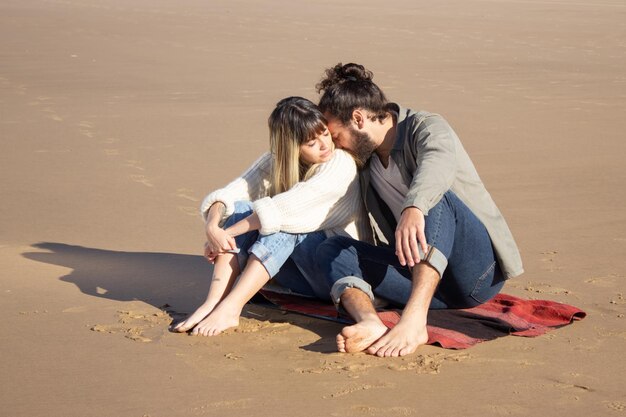 Tenera coppia caucasica in vacanza estiva. Uomo e donna barbuti in abiti casual seduti su una coperta, che si baciano sulla spalla. Concetto di amore, viaggio, tenerezza