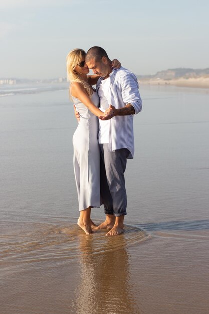 Tenera coppia innamorata in spiaggia. Marito e moglie in abiti casual che si abbracciano e ballano il giorno d'estate. Vacanza, felicità, concetto di relazione