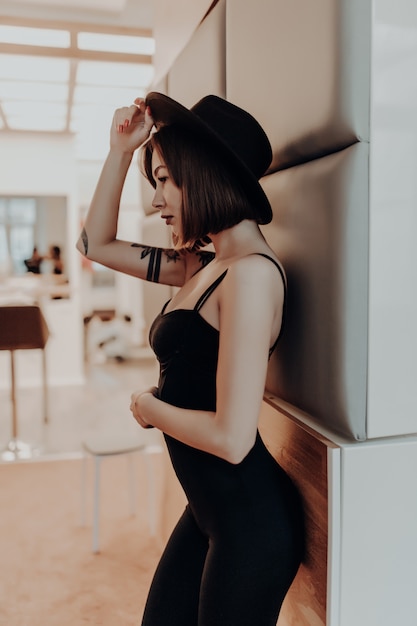 Tenera donna adulta donna bruna in abito nero e cappello in piedi vicino al muro in appartamento di lusso