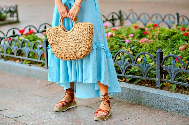 Tenera donna elegante in posa e indossa un maxi abito blu, borsa di paglia e sandali gladiatore