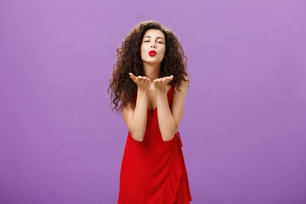 Tenera donna europea affascinante con rossetto rosso in elegante abito da sera che si piega verso la fotocamera piegando le labbra inviando un bacio d'aria alla telecamera che tiene i palmi vicino alla bocca esprimendo amore e passione.