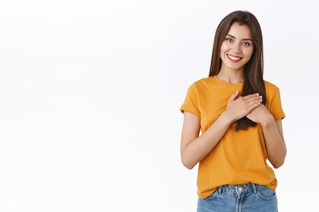 Tenera e affascinante donna caucasica in t-shirt gialla, tenendo le mani premute sul cuore, accarezzando la relazione e l'amore, sorridendo gioiosamente, macchina fotografica dall'aspetto sognante, in piedi soddisfatto sfondo bianco