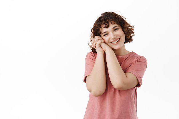 Tenera e bella ragazza riccia, sorridente felice, ammira qualcosa di romantico e carino, appoggiando la testa sulle mani e guardando bella, in piedi in t-shirt su sfondo bianco.