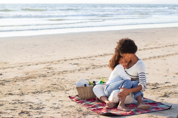 Tenera famiglia afroamericana sul picnic sulla spiaggia. Madre e figlia in abiti casual seduti su una coperta, abbracciati. Famiglia, relax, concetto di natura