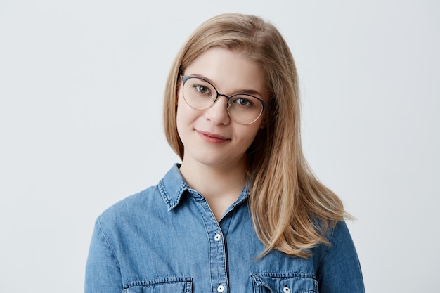 Tenera ragazza bionda con pelle sana che indossa camicia di jeans e occhiali da vista guardando con espressione compiaciuta o pensierosa. Modello caucasico della giovane donna con capelli biondi che posano all'interno