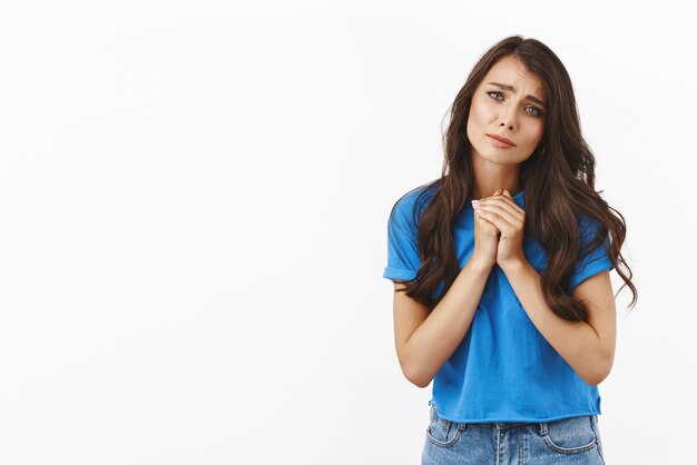 Tenerezza romanticismo concetto Affascinante romantica ragazza bruna carina in maglietta blu tenere le mani insieme accigliata e guardando la telecamera con commovente espressione commossa simpatizzare