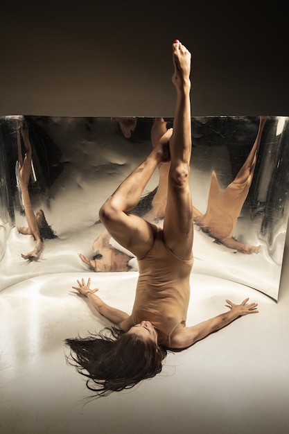 Tenero. Giovane, ballerina moderna alla moda sulla parete marrone con specchio, riflessi di illusione sulla superficie. Magia di flessibilità e movimento. Concetto di arte creativa danza, azione e ispirazione.
