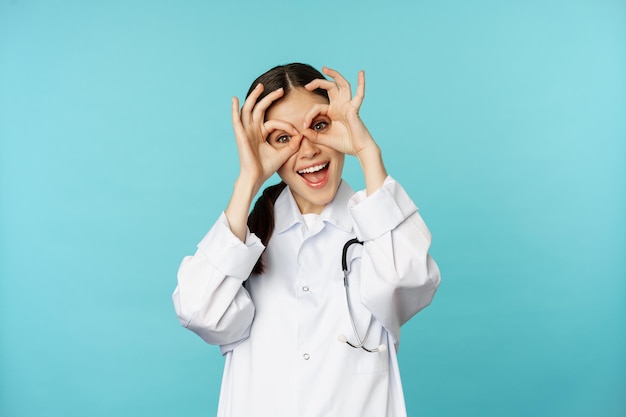 Terapeuta divertente, dottoressa che mostra bene, binocolo zero gesto sugli occhi e sorridente, scherzando e divertendosi, in piedi su sfondo blu.