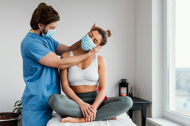 Terapista osteopatico maschio con mascherina medica che controlla i muscoli del collo del paziente femminile