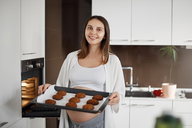 Tesoro, sono a casa. Ritratto di affascinante e premurosa moglie incinta, che tira fuori i biscotti dal forno, tiene la leccarda e sorride ampiamente alla telecamera, aspettando le amiche a casa per discutere la futura maternità.