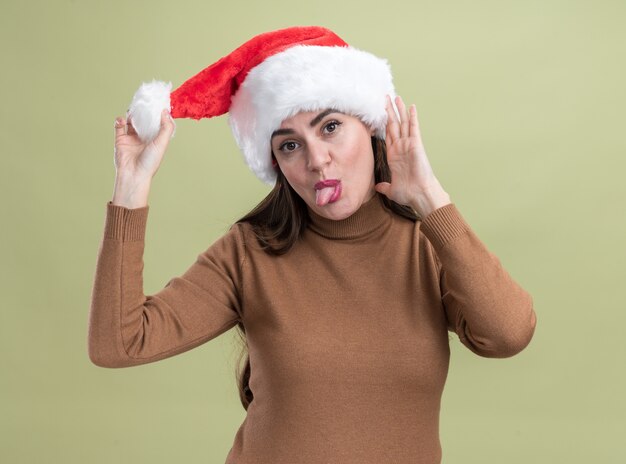 Testa di inclinazione gioiosa giovane bella ragazza che indossa il cappello di Natale che mostra la lingua isolata sulla parete verde oliva