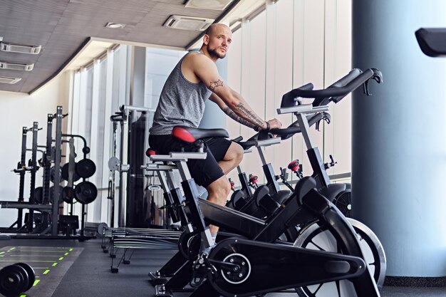 Testa rasata atletica maschio e due femmine di forma fisica esile che si esercitano con la bici del corpo in un club di palestra.