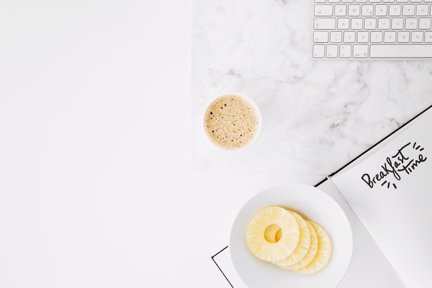 Testo del tempo di colazione su carta con tastiera; fette di ananas e tazza di caffè sulla scrivania