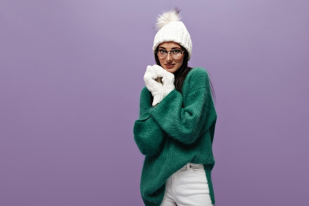Timida donna carina in elegante maglione verde di lana e occhiali posa su sfondo viola Bella ragazza bruna con cappello e guanti guarda nella fotocamera