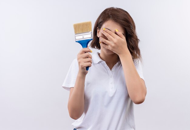 Timida ragazza con i capelli corti che indossa una polo bianca che tiene il pennello che copre gli occhi con la mano