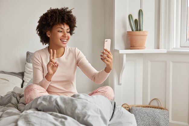 Tiro al coperto di allegra donna dalla pelle scura vestita in pigiama casual, ha una videochiamata mattutina, tiene un moderno telefono cellulare, posa sul letto, sorride ampiamente, mostra il segno di pace sullo schermo. Selfie al mattino