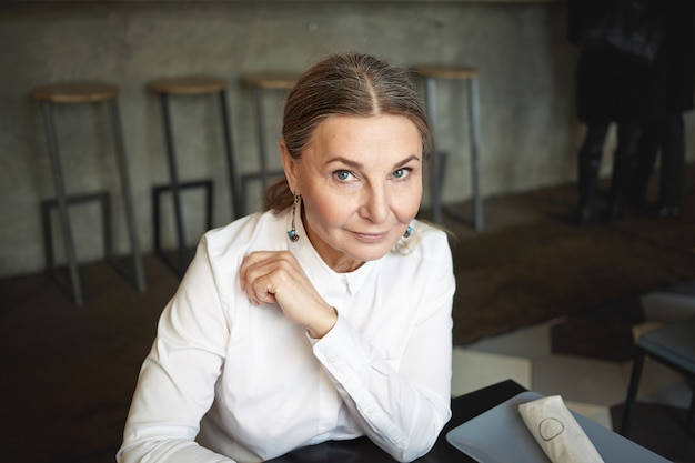 Tiro al coperto di attraente femmina caucasica di mezza età con gli occhi azzurri e un sorriso affascinante pranzando al bar, indossando una camicia bianca e accessori alla moda. Persone, età, stile di vita moderno e svago