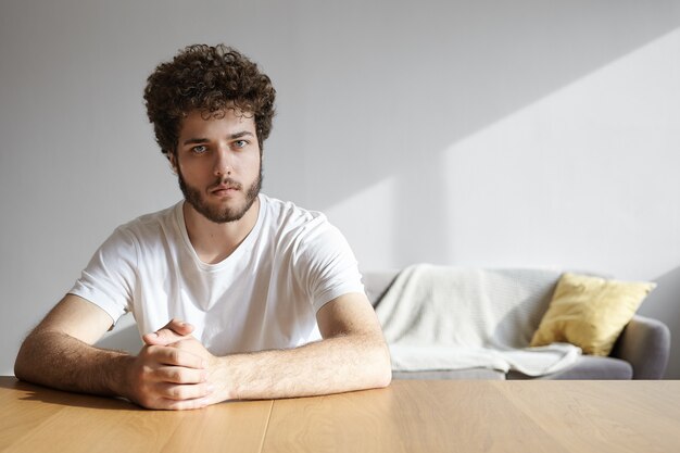 Tiro al coperto di bel ragazzo alla moda giovane hipster con capelli mossi e stoppie seduto al tavolo di legno a casa, tenendo le mani giunte e, andando ad avere una conversazione seria