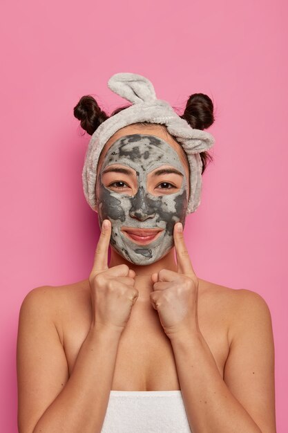 Tiro al coperto di bella donna applica maschera viso peel off detergente nutriente, indica con entrambe le dita anteriori sulle guance, si preoccupa dell'aspetto, ottiene procedure di bellezza nel salone spa, avvolto in un asciugamano