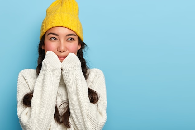 Tiro al coperto di bella donna bruna indossa cappello giallo e maglione bianco, ha un'espressione sognante, distoglie lo sguardo, si erge contro il muro blu