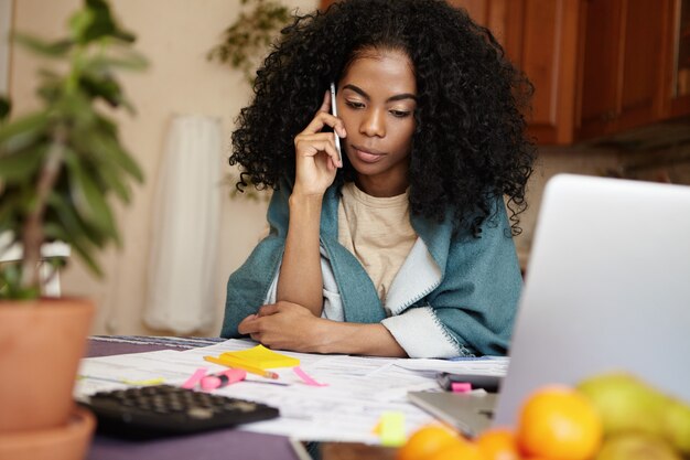 Tiro al coperto di donna disoccupata dalla pelle scura che ha conversazione telefonica con la sua amica, chiedendole soldi per pagare i debiti, seduta al tavolo della cucina con laptop e documenti, calcolando le bollette