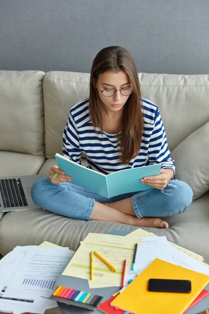 Tiro al coperto di donna freelance focalizzata sul libro, si siede nella posa del loto su un comodo divano, circondata da documenti, lavora da casa, registra le informazioni dalla rete, si prepara per gli esami finali