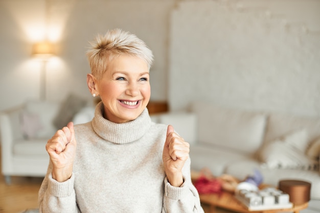 Tiro al coperto di donna matura felicissima alla moda in maglione a collo alto che gode di notizie positive, con un'espressione facciale estatica, ridendo e stringendo i pugni. Concetto di successo e risultati