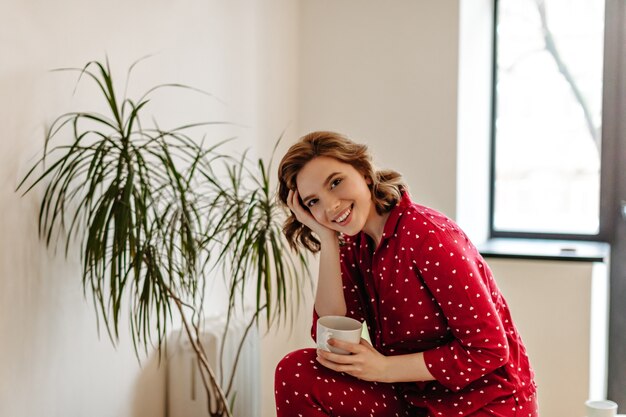 Tiro al coperto di donna sognante in pigiama che beve il tè. Giovane donna caucasica sorridente che tiene tazza di caffè.