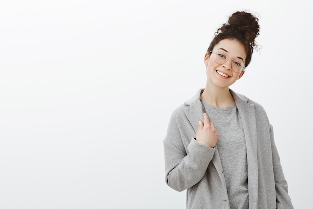 Tiro al coperto di elegante donna dai capelli ricci attraente indipendente in occhiali alla moda e cappotto grigio, toccando i vestiti