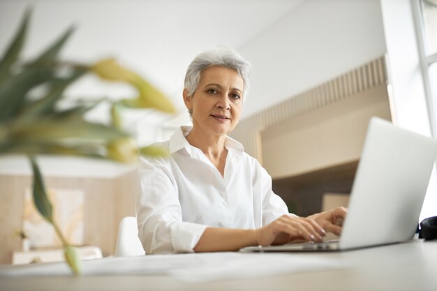 Tiro al coperto di esperto di marketing femminile con esperienza elegante seduto alla scrivania davanti al computer portatile generico aperto