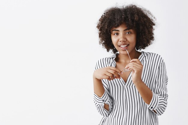 Tiro al coperto di giocosa attraente donna dalla carnagione scura con acconciatura afro che morde il bordo degli occhiali e sorridente con un sorriso gioioso divertito che vuole dire qualcosa ma essere timido sul muro grigio